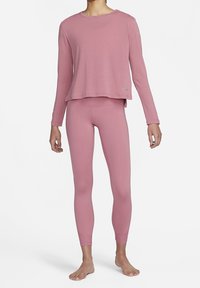 Frau, die ein passendes langärmliges Top und eine enge Leggings in staubigem Rosa trägt, barfuß auf weißem Hintergrund steht.
