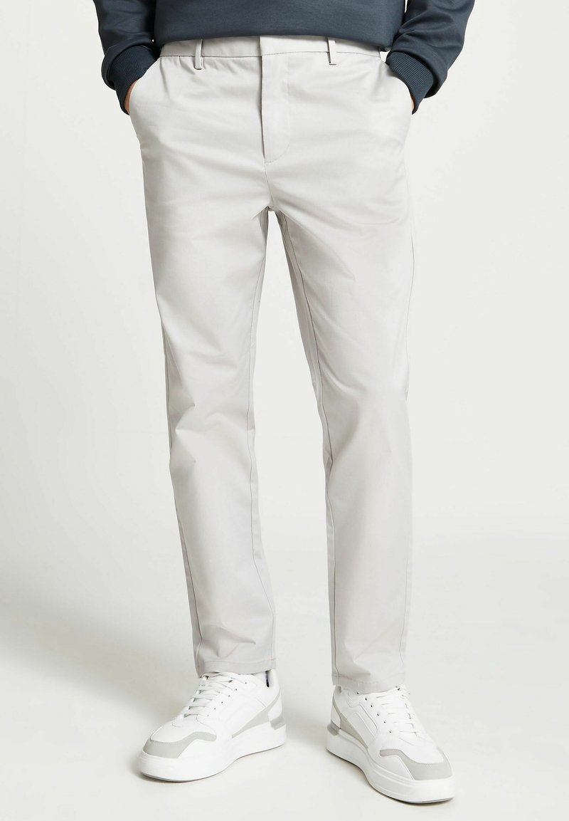 Homme portant un pantalon gris clair ajusté avec les mains dans les poches et des baskets blanches, debout sur un fond blanc uni.