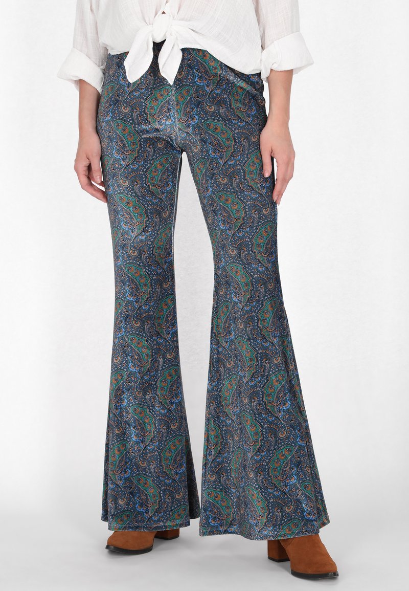Uitlopende broeken in een donkermarineblauw paisleypatroon met groene en bruine accenten. De stof heeft een gladde textuur. Gemaakt bij bruine laarzen met hak.