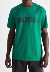 Grøn bomulds T-shirt med korte ærmer, der har et iøjnefaldende sort "BOSS" logo på brystet. Blød tekstur og klassisk pasform.