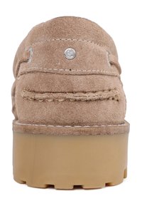 Chaussure en daim marron avec une tige texturée, des coutures visibles et des accents en métal argenté. La semelle est épaisse et transparente.