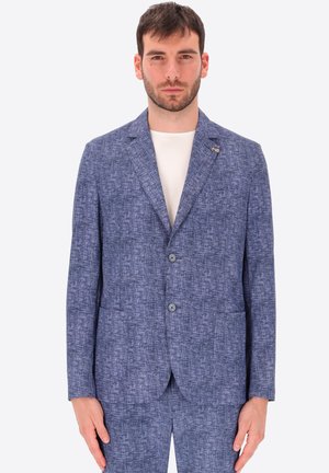 Uomo con capelli corti e scuri che indossa una giacca da completo blu a due bottoni con trama, sopra una maglietta bianca girocollo, in piedi davanti a uno sfondo semplice.