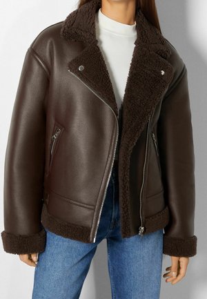 Brązowa kurtka z kożuszkiem, z zamkiem na przodzie, szerokim kołnierzem i mankietami, noszona na białym topie i niebieskich dżinsach przez stojącą osobę.