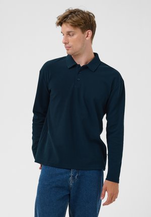 Joven con camiseta polo de manga larga oscura y vaqueros azules, de pie ante un fondo claro y liso, mirando hacia un lado.