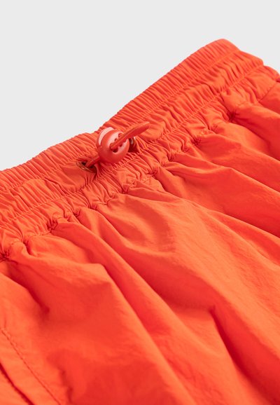 Ceinture élastique orange vif avec un toggle en plastique rond et un cordon sur un tissu léger froncé.