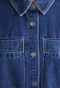 Camisa de mezclilla con cuello clásico, dos bolsillos frontales, botones a presión plateados y costuras en contraste en azul medio. Textura suave.