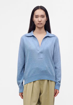 Jeune femme aux longs cheveux foncés portant un pull en maille bleu clair avec col et un pantalon beige plissé sur fond blanc uni.