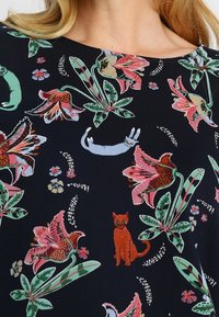 Mörk tyg med färgglada blommönster och lekfulla kattillustrationer i nyanser av grönt, rosa och rött, med varierande former och texturer.