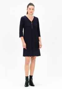 Marineblaues Shiftkleid mit Dreiviertelärmeln, V-Ausschnitt und einem Reißverschlussdetail auf der Vorderseite. Kombiniert mit schwarzen Knöchelboots. Glatte Stofftextur.