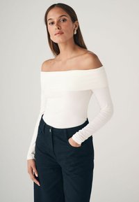 Fehér, váll-levágott, hosszú ujjú bodysuit, testhezálló fazonnal, amelyet magas derekú sötét nadrággal párosítottak. Simaságú anyag, minimális díszítéssel.