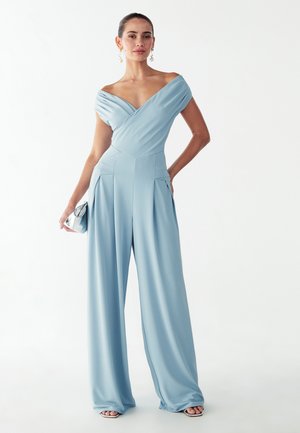Jasnoniebieski kombinezon z odsłoniętymi ramionami, szerokie plisowane nogawki, dopasowane ciało i subtelny połysk. Modelka trzyma srebrną kopertówkę z okrągłymi detalami.