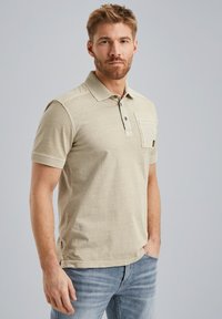 Polo shirt beige clair avec une patte de boutonnage à deux boutons, manches courtes et une poche sur la poitrine. Texture douce et fentes latérales légères au niveau de l'ourlet.