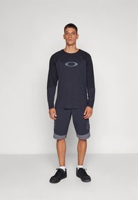 Férfi fekete hosszú ujjú pólóban, ovális logóval és hozzá illő fekete, térdig érő rövidnadrágban áll, egyszínű világosszürke háttér előtt.