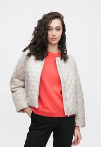 Jasno beżowa pikowana kurtka o krótkim kroju, zapinana na zamek błyskawiczny z przodu. Noszona na jasnoczerwonym swetrze i czarnych spodniach.