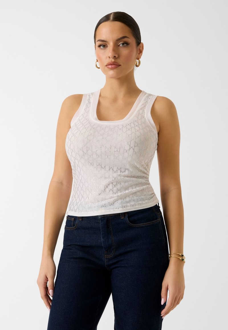 Femme aux cheveux foncés portant un haut sans manches blanc à motifs, un jean bleu foncé, des boucles d'oreilles créoles dorées et des bracelets, debout devant un fond blanc uni.