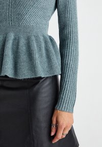 Détail des manches et de la taille d'un pull en maille côtelée teal associé à une jupe en cuir noir, montrant une main avec des bagues en argent.
