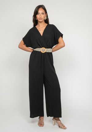 Femme debout, mains sur les hanches, portant une combinaison noire à jambes larges, une ceinture beige à motifs, des boucles d'oreilles dorées et des sandales à talons dorés.