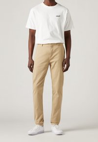 T-shirt bianco a maniche corte con logo nero, abbinato a chinos beige e sneakers bianche. L'outfit presenta una vestibilità rilassata e un design semplice.