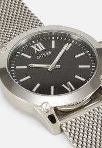 Silbernes Mesh-Armband-Uhr mit einem runden schwarzen gemusterten Zifferblatt, silbernen römischen Ziffern und einer polierten Lünette. Mit Logo und leuchtenden Stundenmarkierungen.