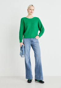Zeleni rebrasti pleteni pulover z širokim izrezom, v kombinaciji s svetlo modrimi širokimi jeans hlačami. Dodatki vključujejo denim torbo in črne čevlje.