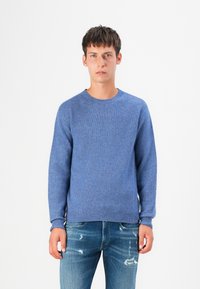Blauwe gebreide trui met een ronde hals en geribbelde manchetten, gecombineerd met versleten blauwe spijkerbroeken. De stof heeft een gestructureerd uiterlijk.