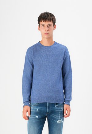 Blå stickad tröja med rundhals, ribbade ärmslut och tillsammans med slitna blå denimjeans. Tyget har en texturerad ytfinish.