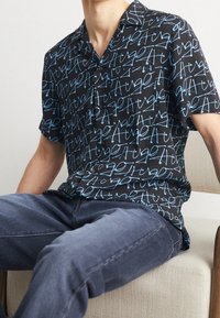 Chemise noire à manches courtes avec un motif graphique bleu répétitif, tissu lisse et col classique.
