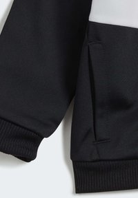 Chaqueta de tela negra con un panel blanco; cuenta con puños acanalados y un bolsillo lateral. Textura suave con detalles de costura limpios.