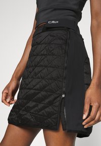 Žena v černé prošívané sukni se zipem na boku a černým pasem se znakem CMP, zobrazená od pasu po polovinu stehna.