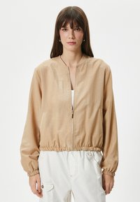 Harmaa vaaleanbeige bomber-takki, jossa on vetoketjukiinnitys, joustava vyötärö ja puffattuja hihoja. Tasainen pinta, jossa on hienovarainen kiilto. Valkoinen toppi alla.