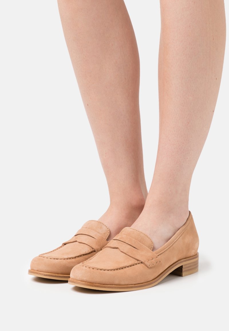 Mocassins en daim beige avec talons empilés bas portés pieds nus sur un fond uni.