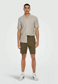 Jovem de pé, a usar uma camisa de botão de manga curta de cor bege claro, calções verde-azeitona, meias brancas e ténis brancos.