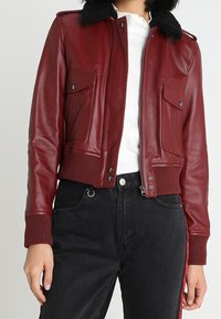 Bordeaux läderbomberjacka med svart pälskrage, frontdragkedja, två bröstfickor, ribbade ärmslut och nederkant. Bärs över en vit skjorta.