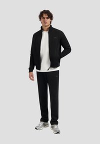 Schwarze Jacke mit Reißverschluss über einem weißen Shirt, kombiniert mit schwarzen Sweathosen. Hat ein einfaches Design und eine glatte Textur, ergänzt durch Sportschuhe.