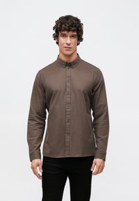 Braunes Langarmhemd mit Knopfleiste am Kragen, mit tailliertem Schnitt, feiner Struktur und Knopfverschluss vorne. Schwarze Hose wird getragen.