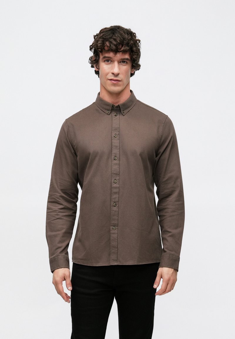 Braunes Langarmhemd mit Knopfleiste am Kragen, mit tailliertem Schnitt, feiner Struktur und Knopfverschluss vorne. Schwarze Hose wird getragen.