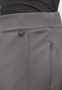 Grå träningsbyxor med ribbad midja, en sidoficka och en liten svart logotyp. Tyget verkar mjukt med en något texturerad yta.