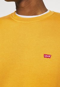 Gele trui van katoen met een geribde ronde halslijn. Heeft een klein rood Levi's-logoplaatje aan de linkerkant.