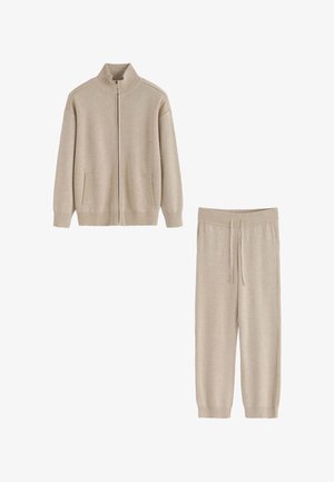 Veste zippée en maille beige avec un col montant, des poches avant, et un pantalon taille élastique assorti avec un cordon de serrage. Texture lisse sur toute la surface.