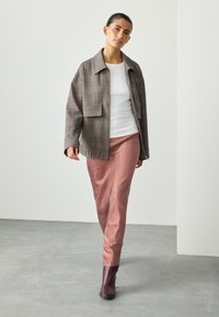 Giacca oversize a quadri in tonalità marroni sopra una maglia aderente bianca, abbinata a pantaloni eleganti in raso rosa e stivaletti marrone scuro.