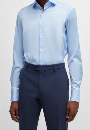 Homme portant une chemise à manches longues bleu clair rentrée dans un pantalon de costume bleu foncé sur un fond uni.