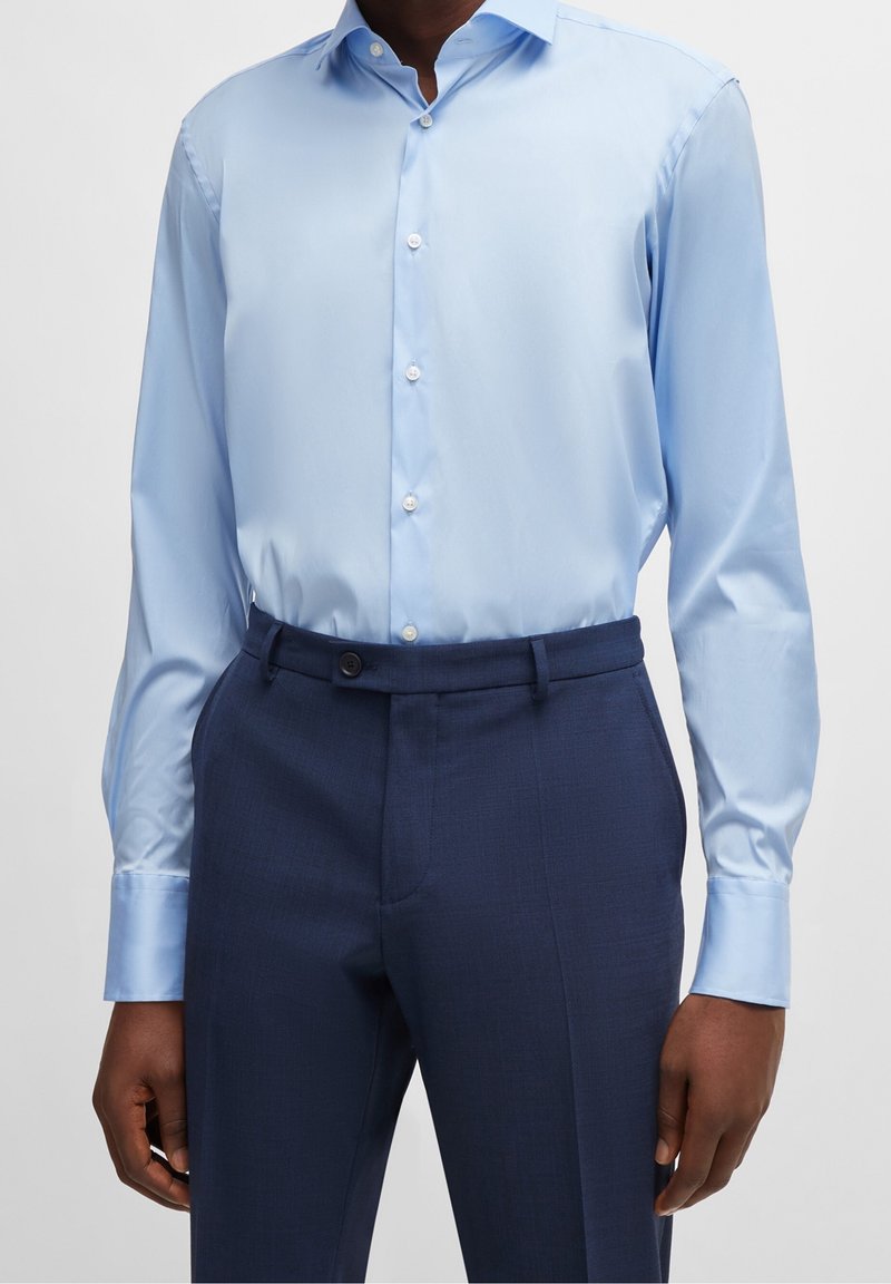 Homme portant une chemise à manches longues bleu clair rentrée dans un pantalon de costume bleu foncé sur un fond uni.