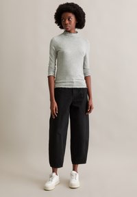 Hellgrünes, geripptes Langarmoberteil mit einem hohen, rüschigen Kragen, kombiniert mit lockeren schwarzen Hosen und weißen Sneakers, vor einem neutralen Hintergrund.