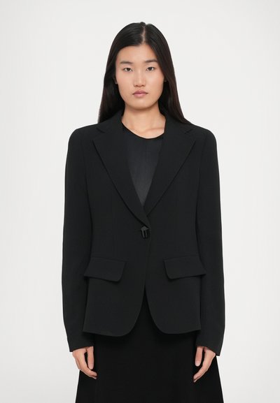 Vrouw met lang steil haar, gekleed in een zwarte blazer met één knoop over een zwart topje, staande tegen een plain witte achtergrond.