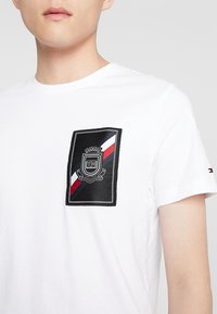 Camiseta de algodón blanca con un parche rectangular negro que presenta un logo de escudo y rayas diagonales rojas y azul marino, cuello redondo, mangas cortas.