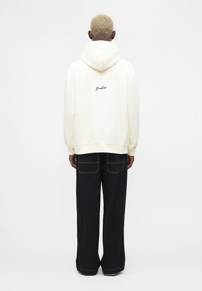 Oseba s kratkimi blond lasmi, oblečena v krem pulover s kapuco in napisom na hrbtu ter črne široke hlače z vidnimi šivi, obrnjena stran.