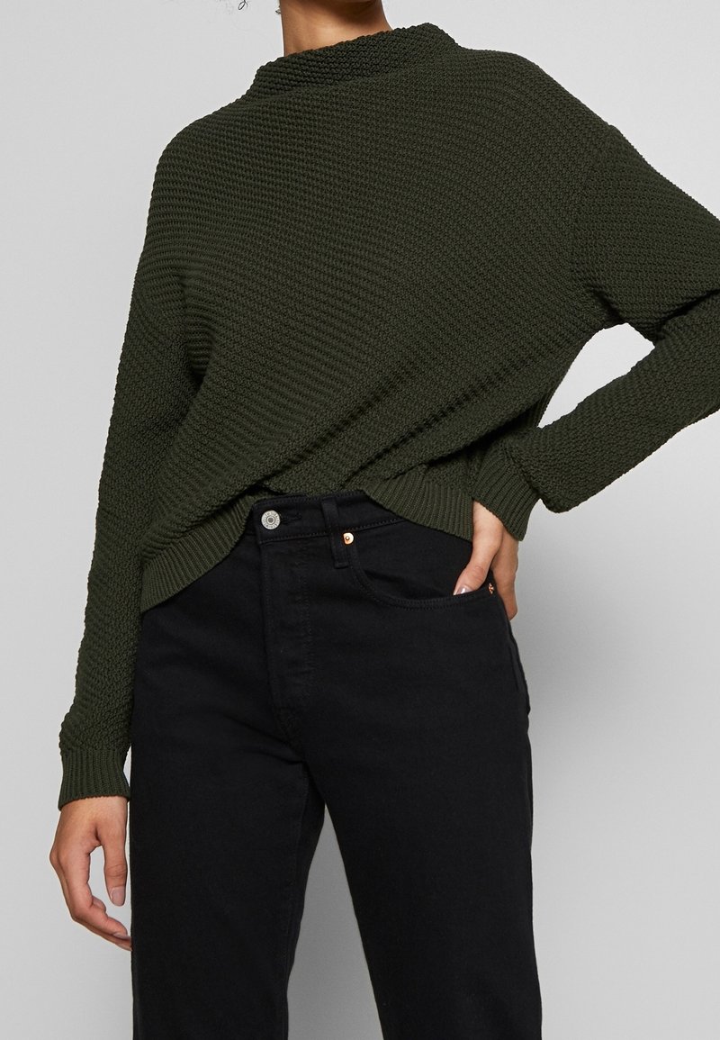 Mörkgrön, texturerad stickad tröja med hög krage, bärs över svarta jeans med knapplås och fickdetaljer.