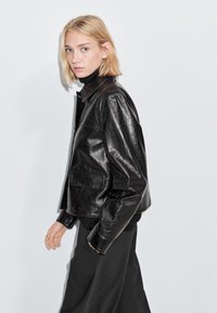 Zwarte leren jas met een glanzende afwerking, gestructureerde schouders en klepzakken. Gestyled met een zwarte coltrui en losse broek.