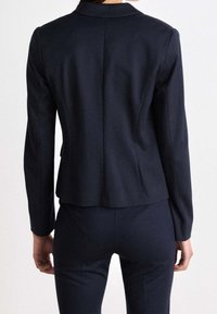 Blazer bleu marine, coupe ajustée, tissu lisse, fente unique à l'arrière, revers crantés, manches longues, coupe cintrée, détails minimalistes au niveau des accessoires.