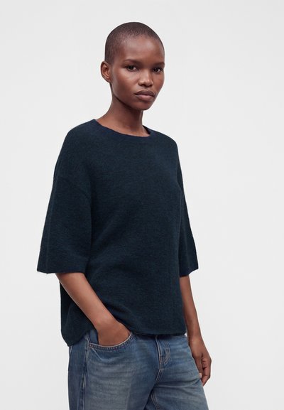Femme aux cheveux courts portant un pull à manches courtes bleu foncé et un jean bleu, debout avec une main dans la poche devant un fond uni.